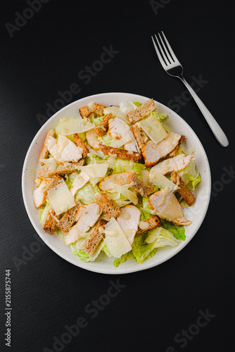 Top view of Caesar salad with grilled chicken, croutons, parmesan shavings and dressing on romaine lettuce, white plate with fork and knife on black wood table, copy space