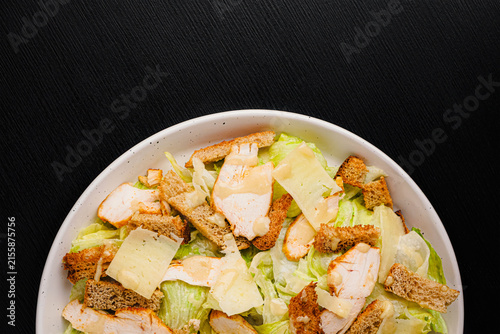 Top view of Caesar salad with grilled chicken, croutons, parmesan shavings and dressing on romaine lettuce, white plate on black wood table, copy space