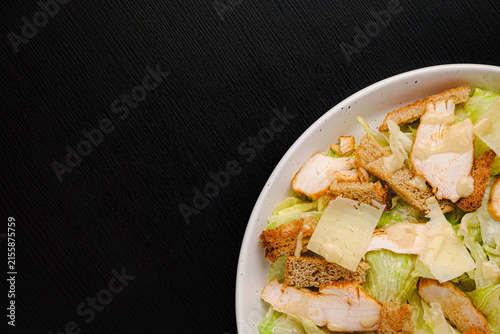 Top view of Caesar salad with grilled chicken, croutons, parmesan shavings and dressing on romaine lettuce, white plate on black wood table, copy space