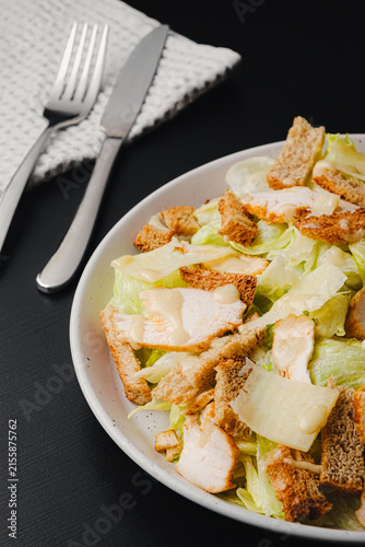 Top view of Caesar salad with grilled chicken, croutons, parmesan shavings and dressing on romaine lettuce, white plate with fork and knife on black wood table, copy space