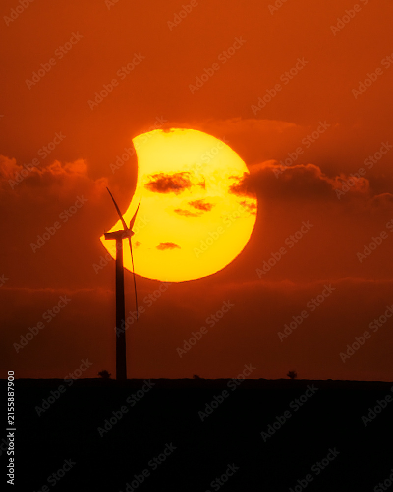 Fototapeta premium Partial solar eclipse behind a wind turbine silhouette during sunset, combining renewable energy concept with astronomical phenomenon