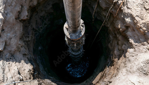 Submersible pump descends into deep borehole with metal pipe, wire rope. Industrial mechanism installs well for water extraction inside dark circular shaft. Technical engineering equipment provides