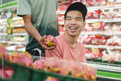 Happy young Asian man in wheelchair choosing fresh dragon fruit with father in supermarket. Disabled male customer enjoying grocery shopping, family support, accessibility and inclusion concept.
