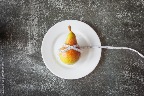 Fotografie Ripe pear wrapped in measuring tape on a white plate against a dark gray studio