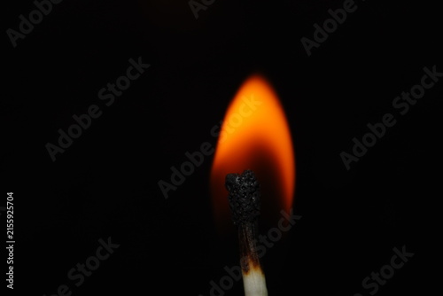 Close-up macro photograph of a burning matchstick with a glowing orange flame isolated against a deep black background.