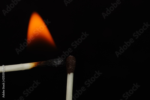 Macro photograph showing two matchsticks positioned together beneath a glowing orange flame on a black background.
