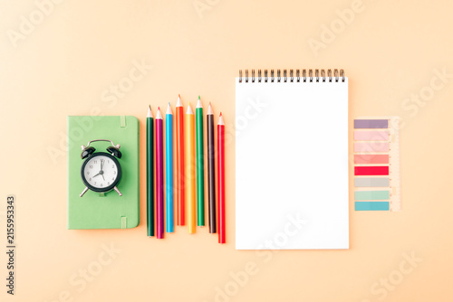 Colorful stationery with pencils, notebook, ruler and alarm clock arranged on a beige background with copy space. Top view, flat lay