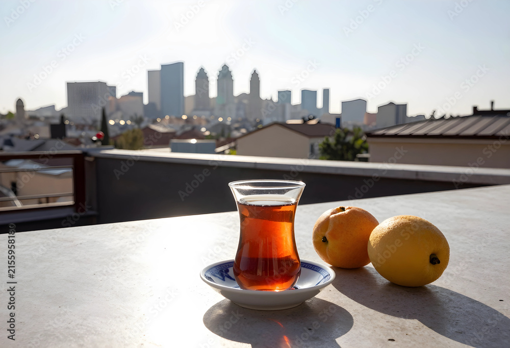 Fototapeta premium Turkish tea in a traditional glass with fresh apricots on a rooftop terrace overlooking a city skyline, the blurred background features a modern city skyline with tall buildings under a clear sky