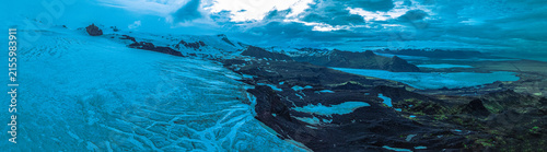 Panoramic aerial view of Vatnajökull Glacier flowing into Iceland's volcanic highlands with rugged lava fields, glacial lagoons and dramatic mountain scenery beneath stormy skies.