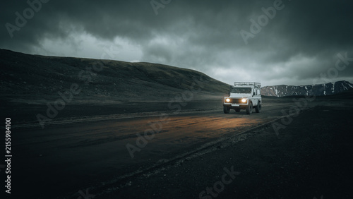 An off-road vehicle drives across the remote volcanic Highlands of Iceland beneath dark storm clouds, with headlights illuminating the rugged track through an untouched wilderness.