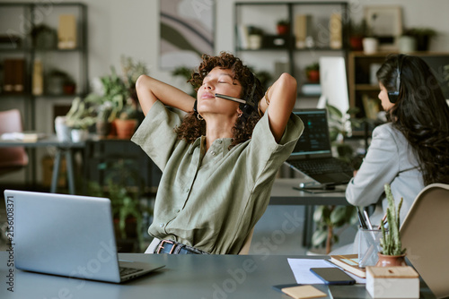 Diverse young adult female call center operators working in office, one stretching with closed eyes. Image represents mental wellbeing and stress relief in corporate communication