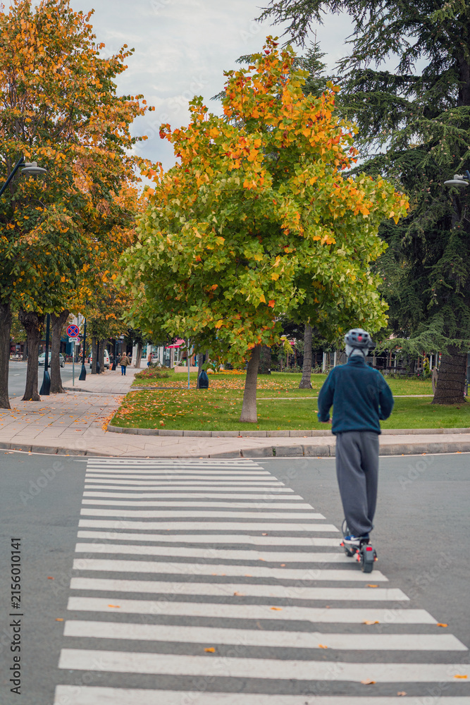 Fototapeta premium Person Riding Electric Scooter Across Urban Crosswalk in Autumn. Young man riding an electric scooter, pedestrian crossing, autumn city street. Urban micromobility, sustainable personal transportation