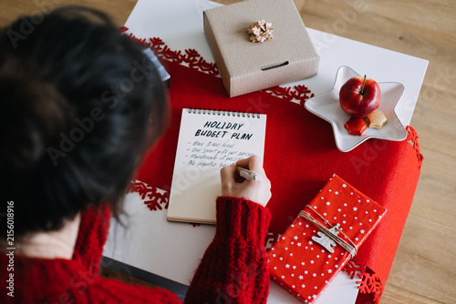 Woman writes in notebook with HOLIDAY BUDGET PLAN on table beside gifts and COZY CHRISTMAS sign. Saving money for christmas, financial goal setting holiday, christmas expense management