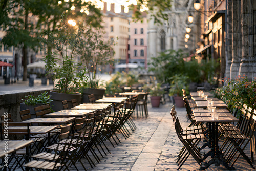 Empty outdoor cafe tables and chairs on a city sidewalk bathed in warm golden hour light, creating long shadows. A tranquil urban scene.