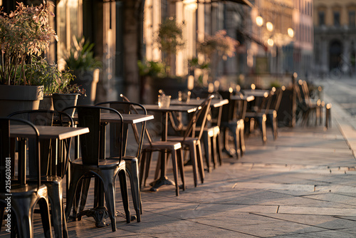 Empty outdoor cafe tables and chairs on a city sidewalk bathed in warm golden hour light, creating long shadows. A tranquil urban scene.