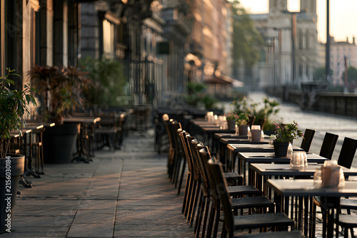 Empty outdoor cafe tables and chairs on a city sidewalk bathed in warm golden hour light, creating long shadows. A tranquil urban scene.