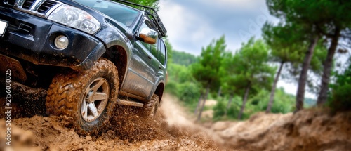 Dark off road vehicle moves through deep mud, wheels spraying water and mud in intense action against a soft focus forest background