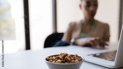 Bowl of snacks beside a laptop with a woman in the background, illustrating food noise, distracted eating and habitual cravings during work or screen time