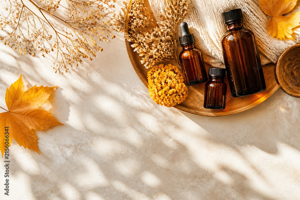 Naklejka premium A flat lay composition featuring amber essential oil bottles, dried flowers and foliage, and a fallen autumn leaf on a light surface.