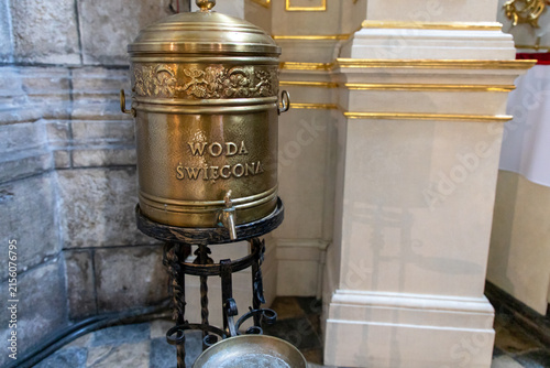 Foto A brass church holy water dispenser featuring an integrated tap and the Polish inscription WODA ŚWIĘCONA mounted on a decorative wrought-iron stand