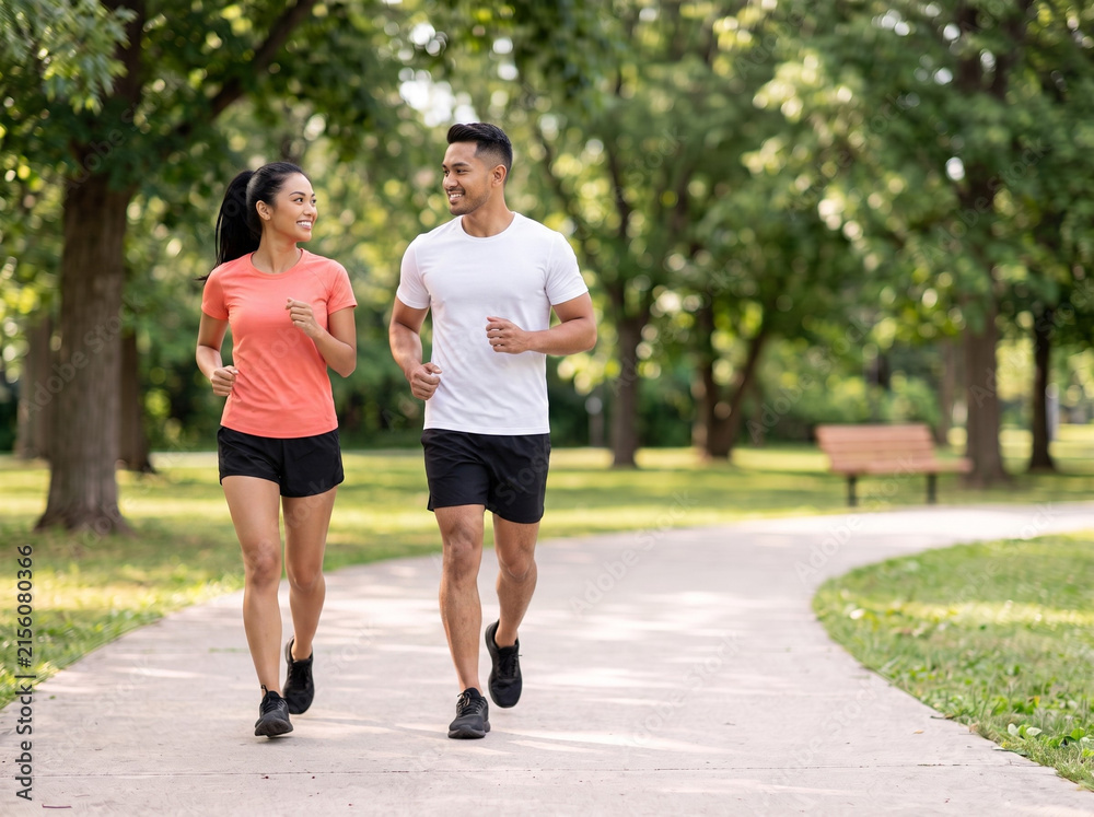 Fototapeta premium Young Southeast Asian couple jogging together in a green park for fitness wellness heart health relationship goals healthy lifestyle and outdoor exercise marketing