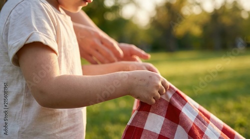 A child grips a red and white checkered picnic blanket as another person arranges items for a meal outdoors in a green park