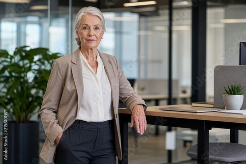 Confident senior businesswoman in a suit standing by a desk in a modern office, concept of successful mature professional, experienced leadership and active career in later life
