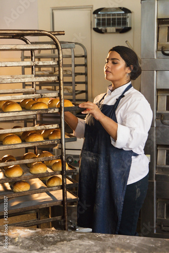 Woman in chef coat arranging trays of golden rolls on metal rack at bakery, copy space
