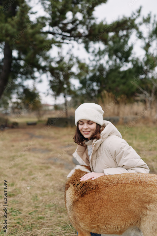Naklejka premium Girl in a warm beige coat and white beanie outdoors, smiling as she pats a fluffy alpaca in a grassy field on a cool day, sharing a wholesome nature moment