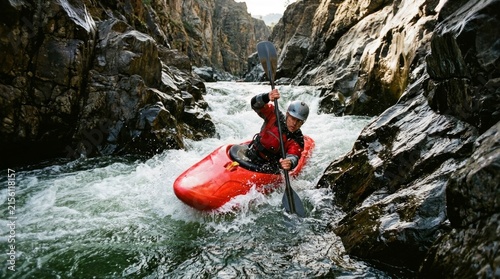 A person paddles a red kayak through white water between rocky cliffs.
