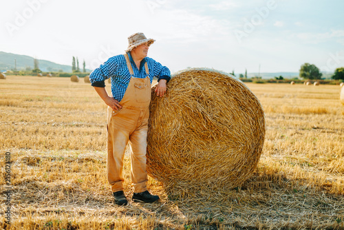 Full body view of older male farmer resting against a hay bale in summer.