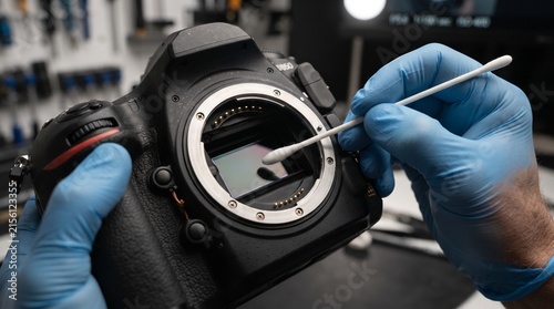 A close-up shot shows a pair of gloved hands carefully cleaning a digital camera sensor with a cotton swab in a dimly lit workshop, highlighting the delicate process of camera maintenance.