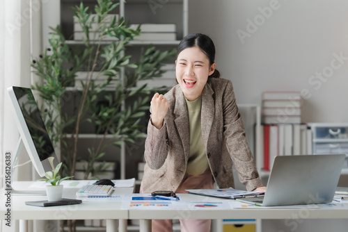 Canvas Print Excited Businesswoman Celebrating Success: A young, energetic businesswoman pumps her fist in triumph while working at her modern office desk, surrounded by success and motivation