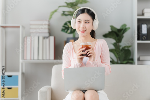 Canvastavla Relaxing Melody: A young woman enjoys a refreshing iced coffee while listening to music on her headphones, laptop on her lap, in a cozy and bright living room