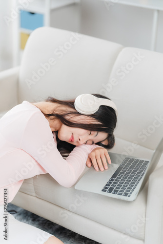 Overworked and Exhausted: A young woman in a light pink sweater, wearing white headphones, sleeps on a sofa with her head on a laptop, indicating exhaustion and overwork.