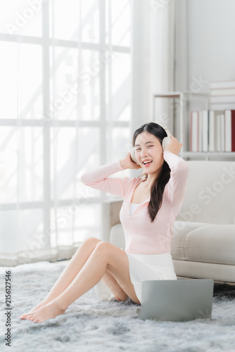 Принти на полотні Young Woman Listening to Music with Laptop: A young woman sits on a rug, wearing headphones and looking at a laptop, with a happy expression, enjoying music in a bright room