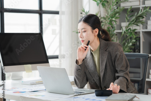 Focused Professional at Desk: A focused young woman in professional attire works diligently at her desk, surrounded by office supplies, a computer, and paperwork, in a bright and modern workspace.
