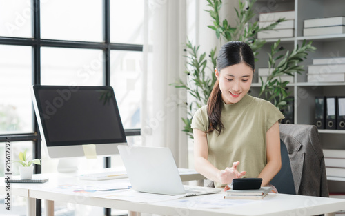 Cuadro en lienzo A Young Woman Calculating at her Desk: A focused young woman, seated at a modern office desk, diligently uses a calculator, surrounded by a computer, laptop, and files