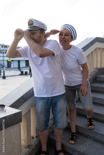 Middle aged couple in captain and sailor hats looking away on river station embankment