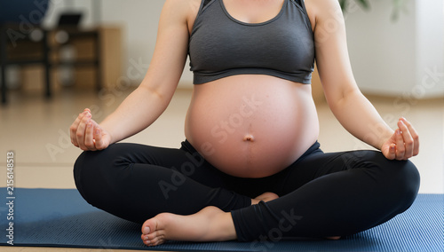Close-up of young pregnant woman sitting on exercise mat and meditating during her training
