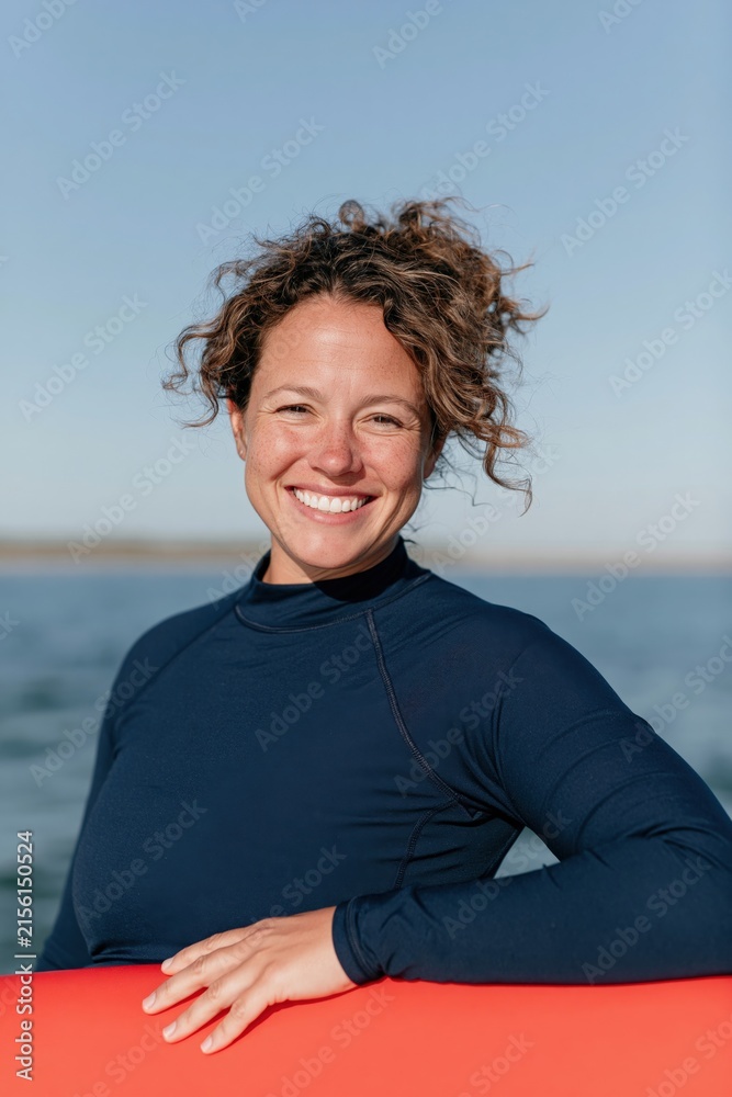 Fototapeta premium Happy woman in wetsuit holding red surfboard by the sea for outdoor sports portrait and active lifestyle banner