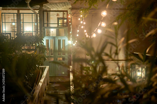 Night courtyard with wooden deck and glass doors. String of warm lights, blurred plants in the foreground. Cozy cafe or veranda atmosphere.