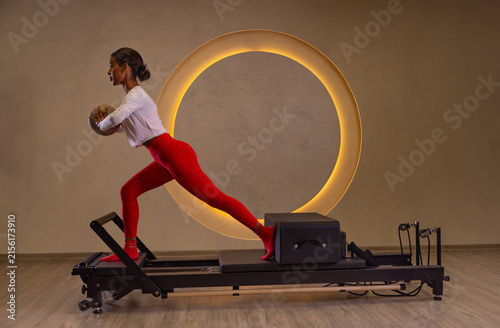 Woman exercising on a Pilates Reformer in a modern wellness studio, with a glowing crescent moon design in the background. Fitness, strength, flexibility, and mindful movement.