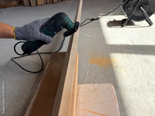 Worker sanding the edge of a wooden plank with an electric detail sander. A construction or renovation site