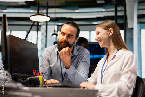Jolly system administrator mentors a junior developer at an office desk, teaching coding best practices and guiding the female intern through project tasks at a technology startup.