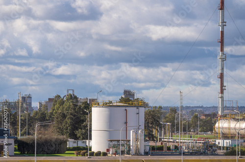 Large white cylindrical industrial storage tank surrounded by green trees, metal infrastructure, light poles, and a tall lattice communication mast against a cloudy blue sky