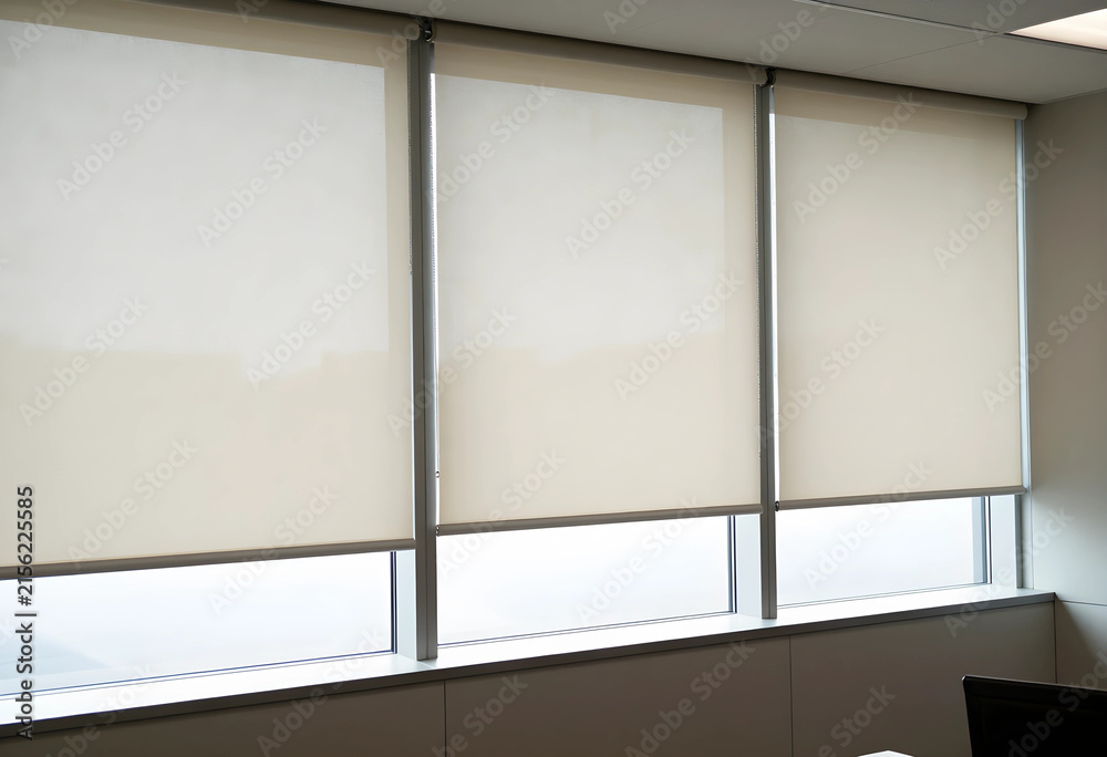 Fototapeta premium Modern office window with three beige roller blinds partially lowered, providing privacy and light control in a contemporary interior