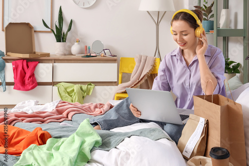 Young woman in headphones using a laptop on a bed with Scattered clothes and ...