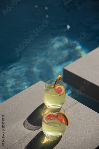 Two stemless glasses sitting on light-gray pool steps, green drink, citrus on rims, water shining