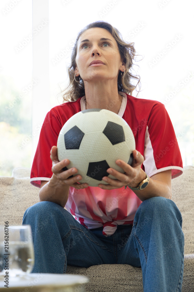Fototapeta premium Woman wearing red-white soccer jersey sitting on sofa holding black-white ball by window watch seen
